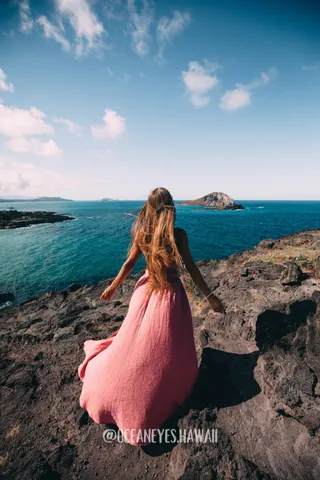 Woman in a pink dress on a cliff above the turquoise ocean in Hawaii