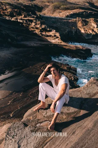 Man in white sitting on lava rocks by the ocean in Hawaii