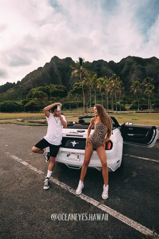 Couple posing by a white convertible with green mountains in Hawaii